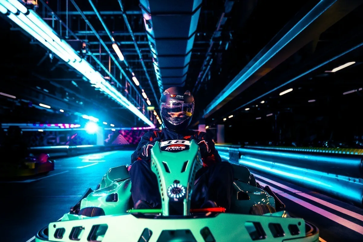A go-kart racer in a helmet on an indoor track with dramatic blue lighting and neon accents creating a futuristic atmosphere.