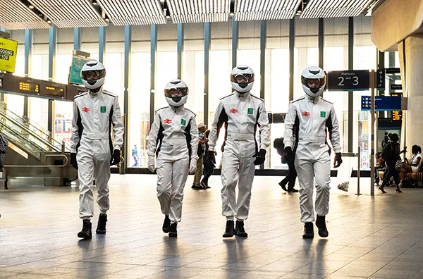 4 people dressed in white motor racing jumpsuits and wearing white racing helmets, walking side by side towards the camera  in the hall of a large railway station.