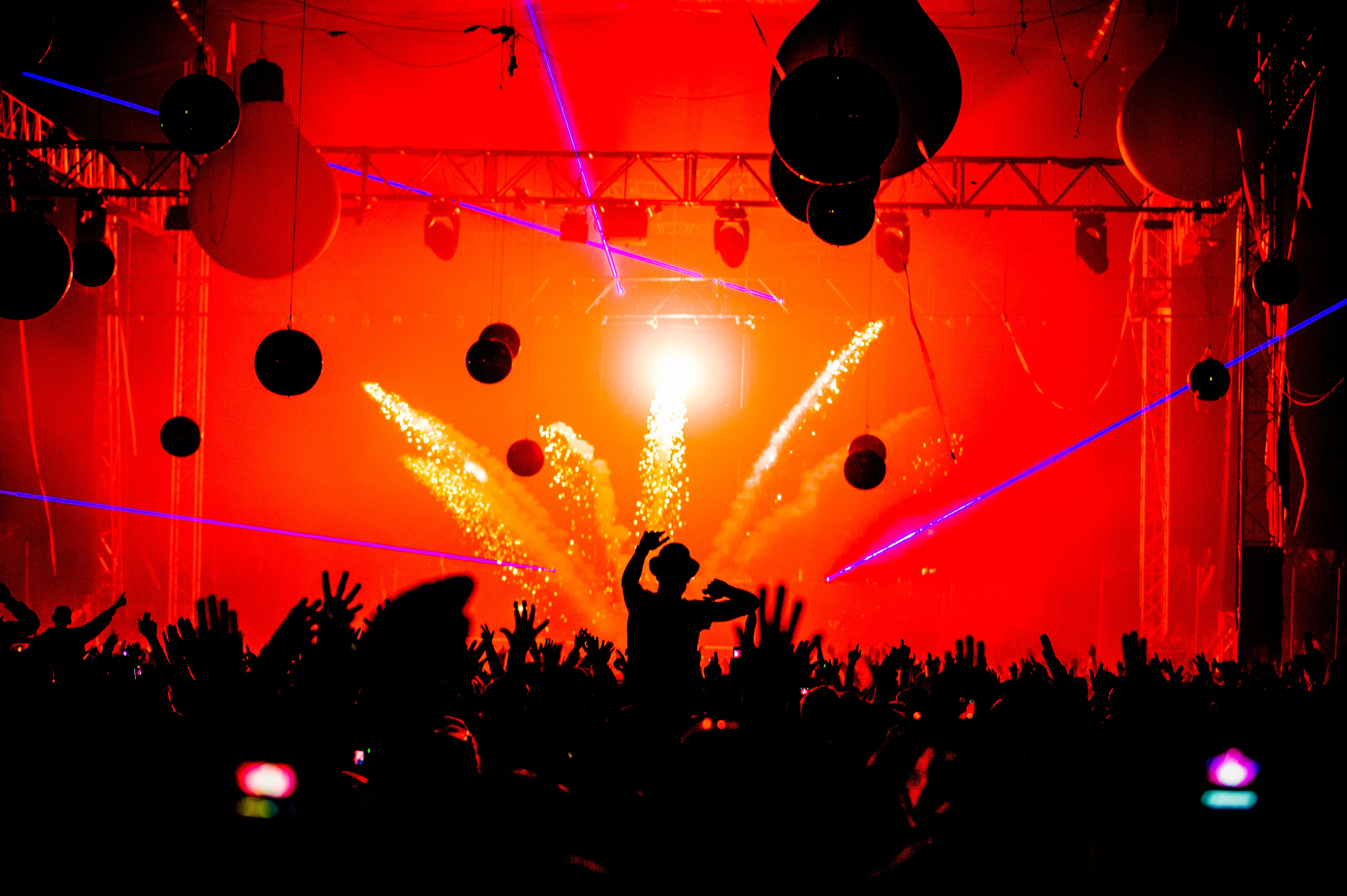 Festival goers take in the pyrotechnics and bright show. Silhouettes of festival goers can be seen in the foreground, with a bright stage and sparks in background.