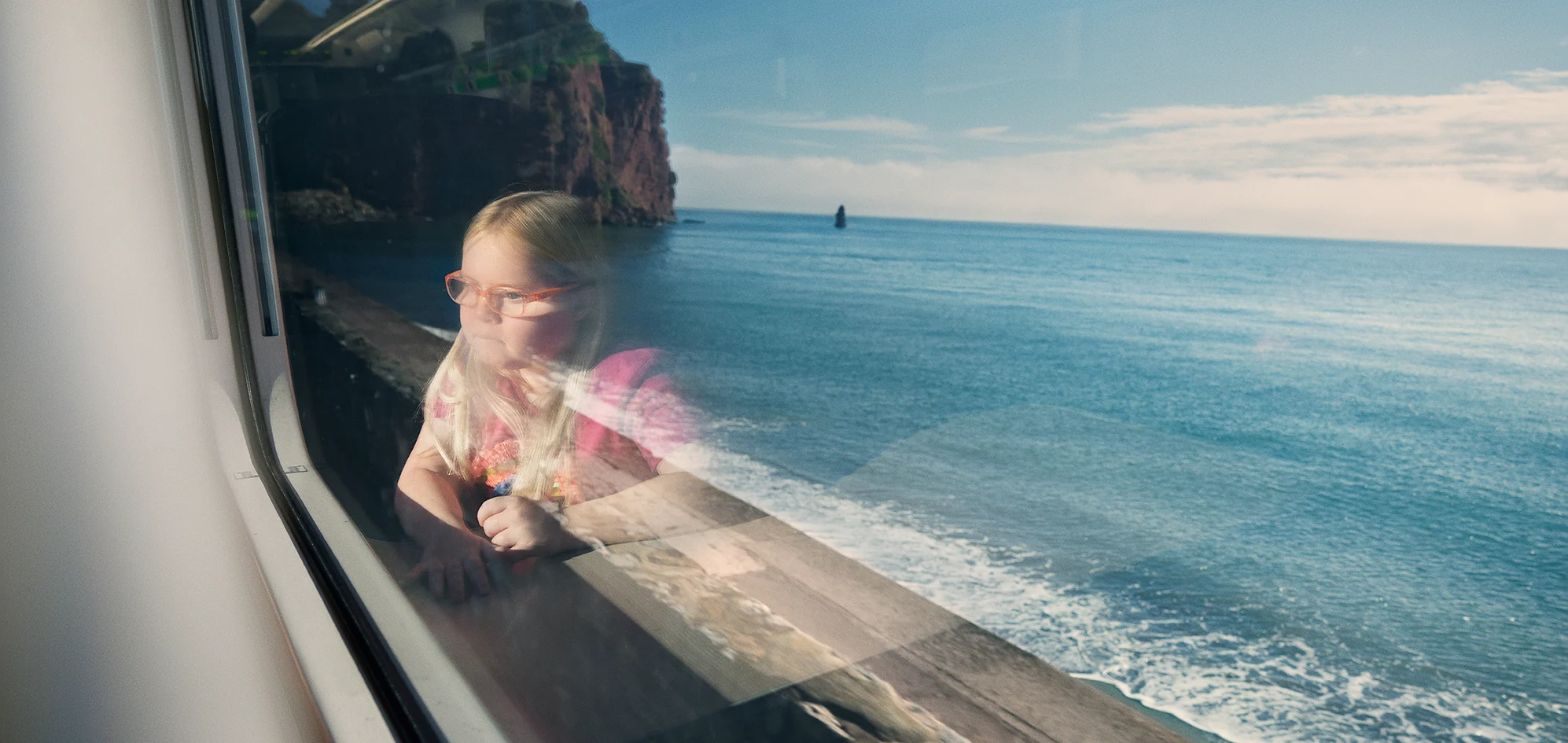 A young girl with blonde hair and red glasses reflected in a train window, sitting at a table in a train carriage and looking out over a beach and the sea.