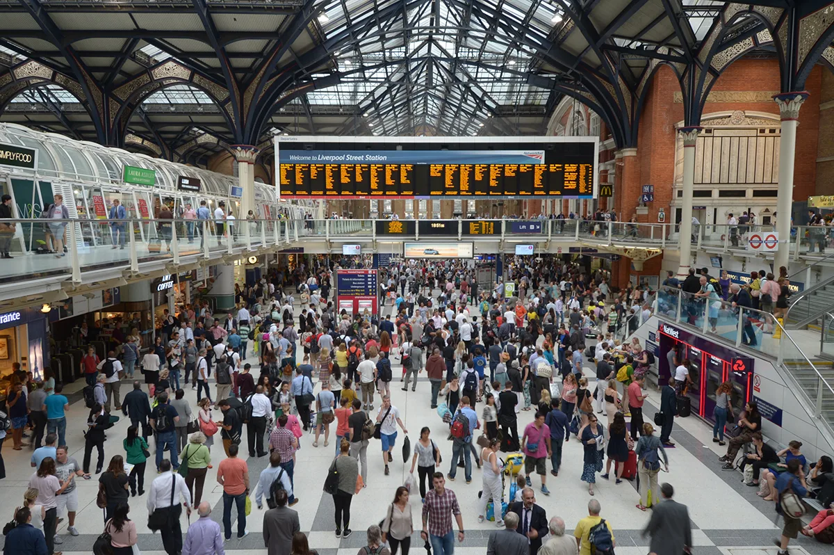 Looking down over the busy concourse at London Liverpool Street station, with a large departure board in the distance and a glass and metal roof visible above. 