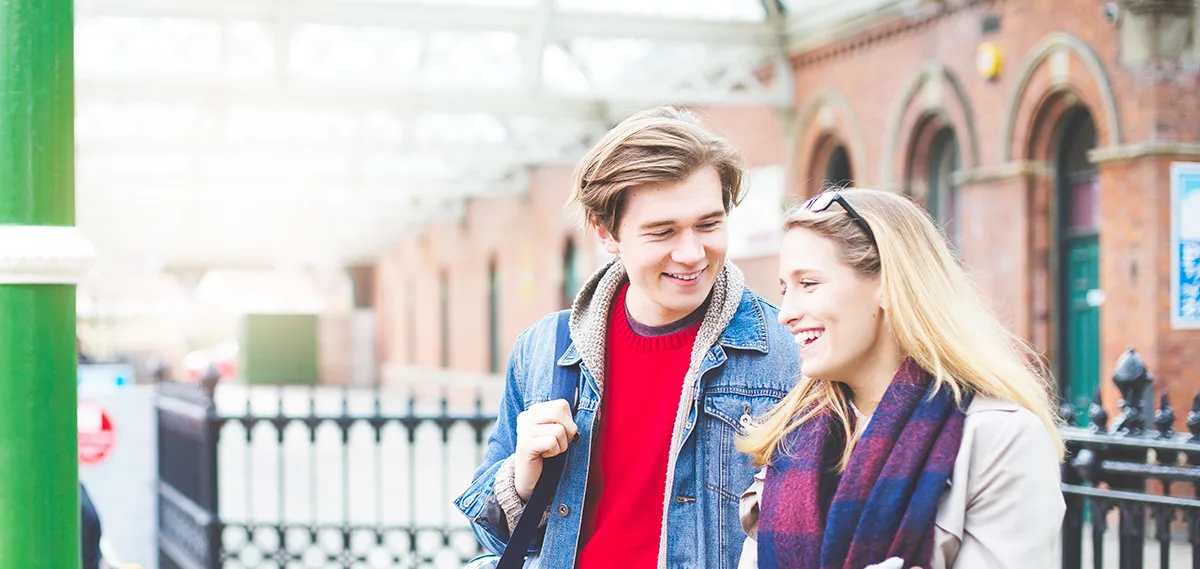 A young white man carrying a shoulder bag and a young white woman wearing gloves and a scarf, smiling and walking at a station.