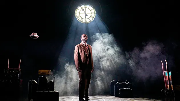A Black man in a brown suit standing beneath a spotlight in front of a clock, part of the cast of the play Harry Potter and the Cursed Child