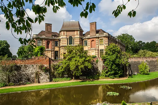 Historic manor house with brick and stone façade surrounded by gardens, stone walls and a reflective pond under blue sky.