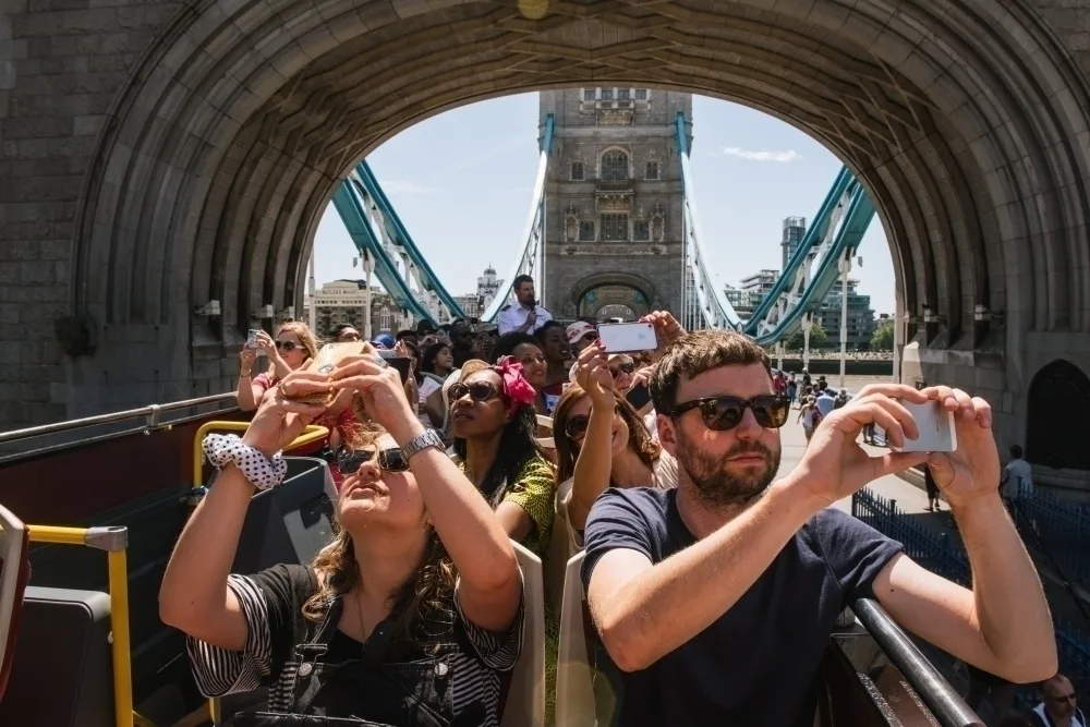 Passengers on an open-top bus taking photos of Tower Bridge in London through a stone archway on a sunny day.