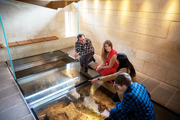 A group of people kneeling on a glass floor in a historical building and looking through to the relics below. 