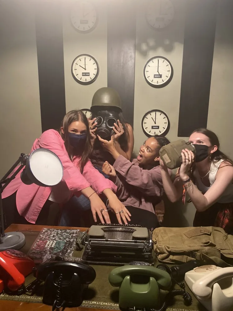 A group of people in an escape room, some wearing helmets and gas masks, with clocks on the wall and old-fashioned telephones on a table.