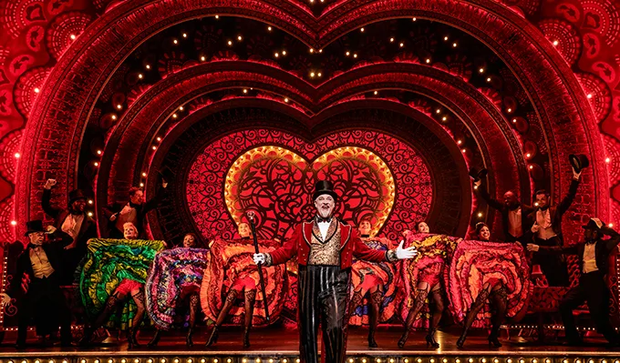 A lavish theatre stage with a giant red heart backdrop, can-can dancing girls and an MC wearing a top hat and red jacket and carrying a cane.