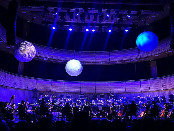 An orchestra on stage bathed in purple light with 3 large globes suspended above. 