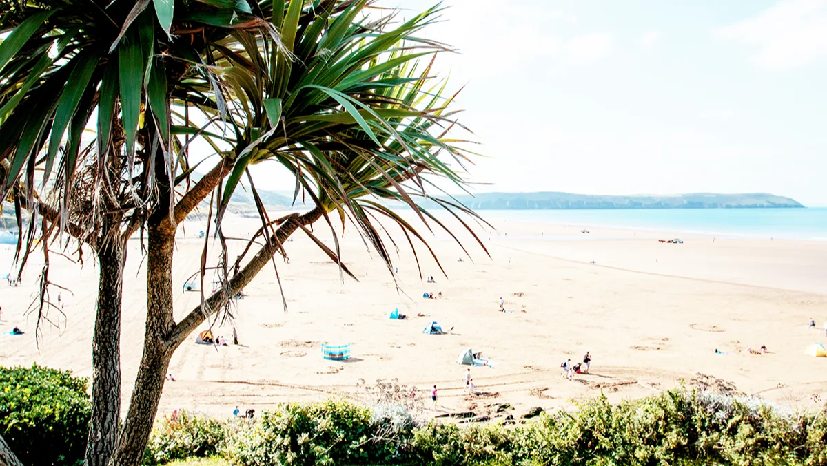 View over a sandy beach dotted with people, with bright blue sea to the right and green hills in the distance, on a sunny day.