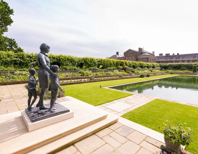 A statue of Princess Diana looking out over a formal garden with a pond, with Kensington Palace in the background. 