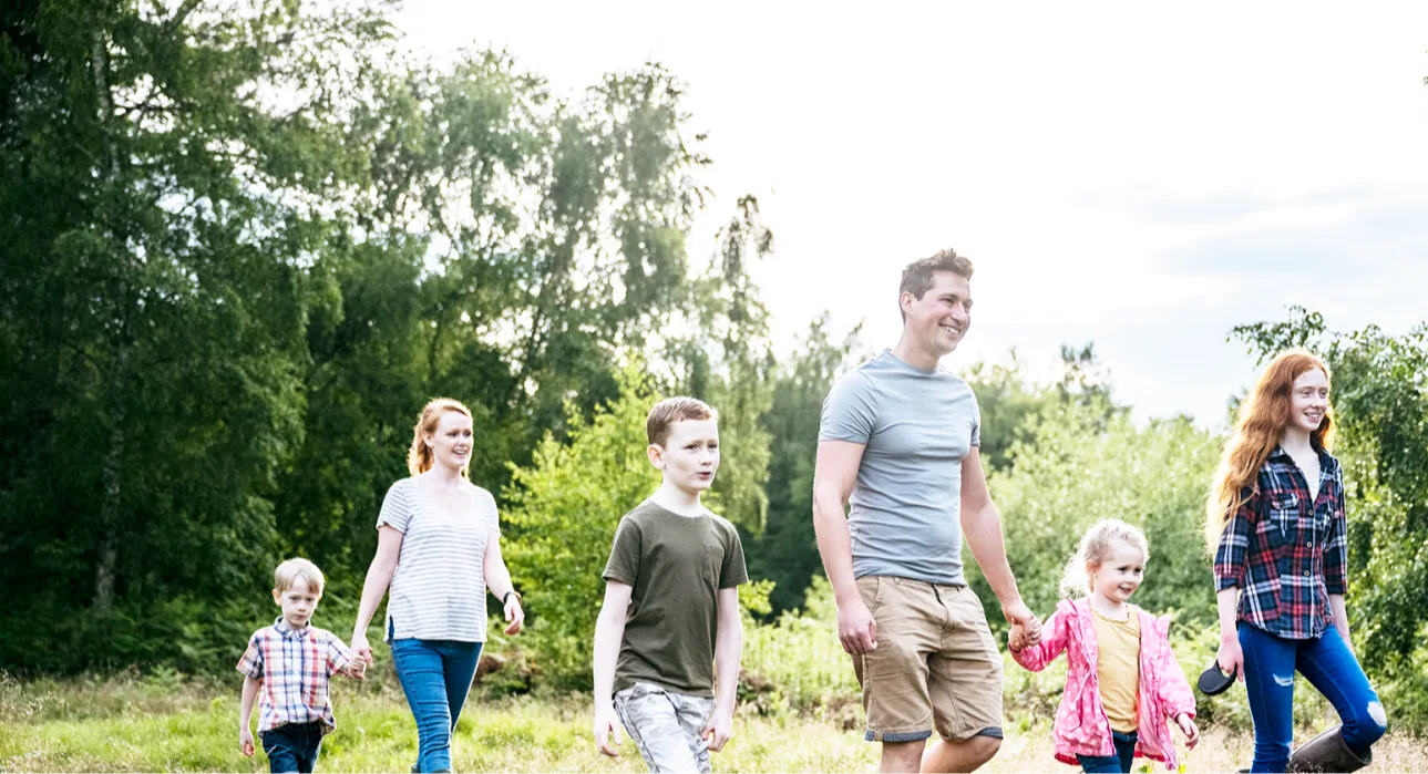 A white family (a man, a woman and 4 children) walking through a woodland area on a sunny day.