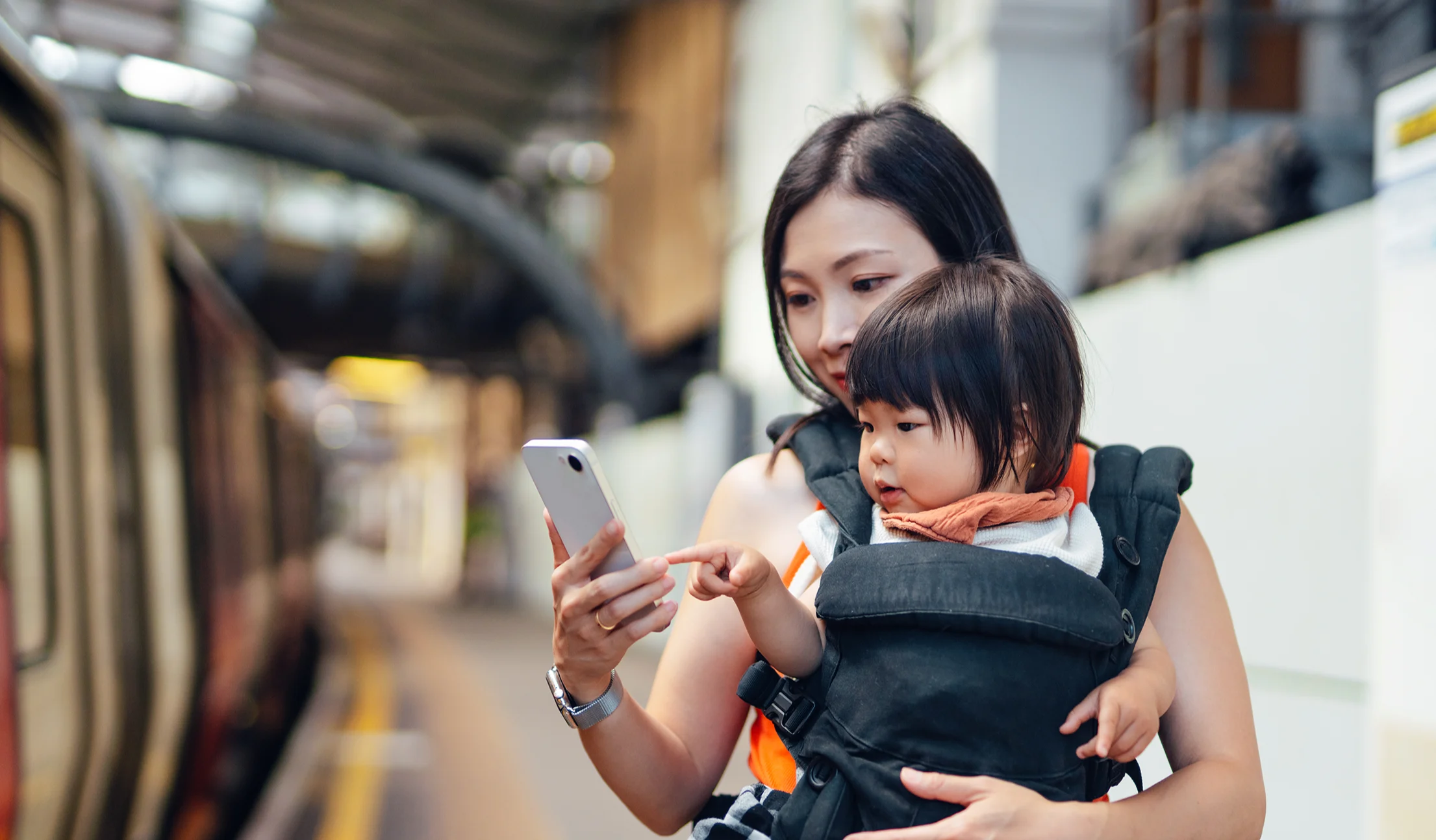 A woman wearing a baby in a front-facing carrier checks her mobile phone on a station platform.