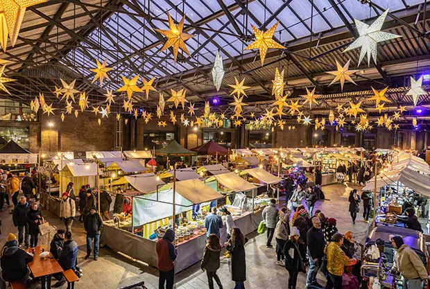 An indoor market with illuminated Christmas star decorations hanging from the high ceiling and people walking around a number of market stalls. 