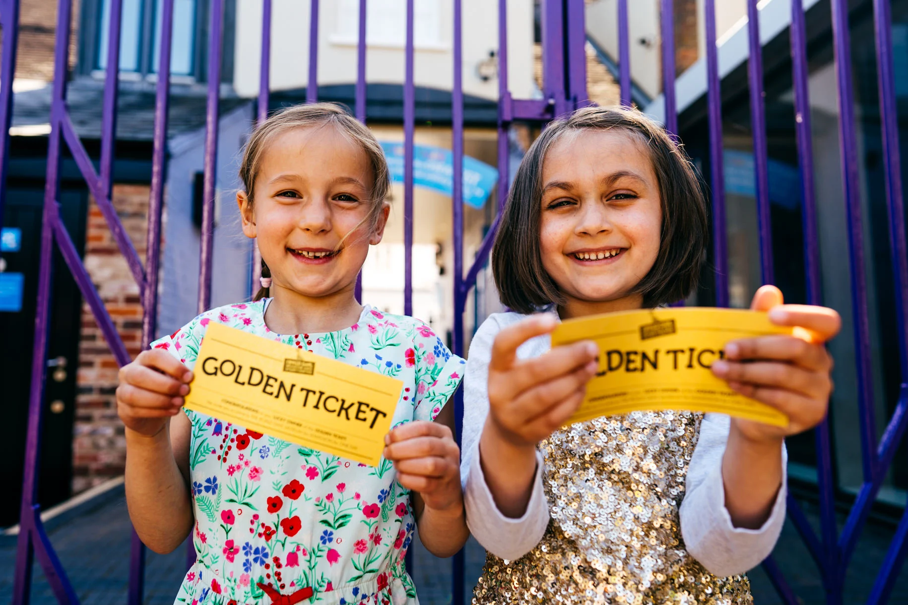 Two smiling children holding yellow 'golden tickets', standing in front of purple railings.