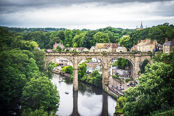 A stone railway viaduct crossing a river with a small town on the banks and trees and hills surrounding it. 