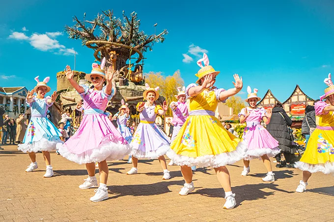 A group of white women doing a synchronised dance while dressed in pastel coloured dresses and straw hats with rabbit ears