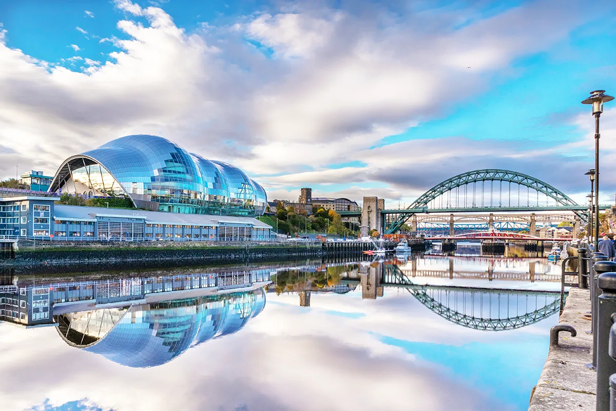 A curved glass-paned building and a bridge on the banks of a river reflected in the water below. 