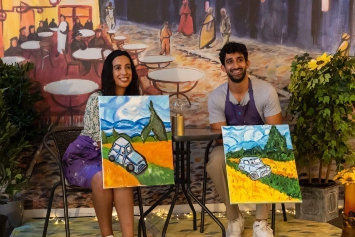A South Asian woman and man sitting in chairs at a cafe style table, they are both holding paintings of a car in a field. 