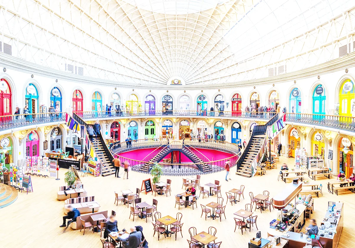 The interior of a circular shopping centre building with cafe tables on a mezzanine floor and a variety of brightly coloured doors on 2 levels. 
