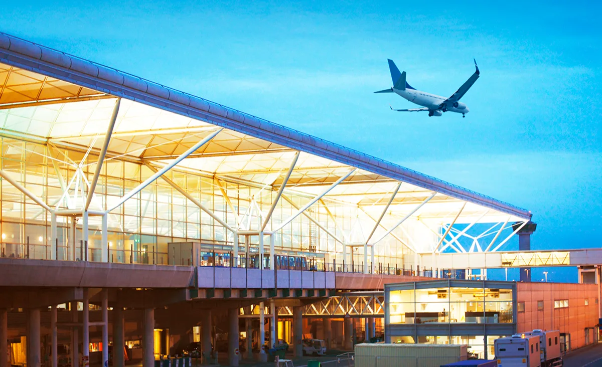 Passenger aircraft descending over Stansted airport terminal with illuminated glass facade against a blue evening sky.
