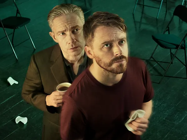 A middle aged white man stands close behind a younger white man with a beard, both holding paper coffee cups. 