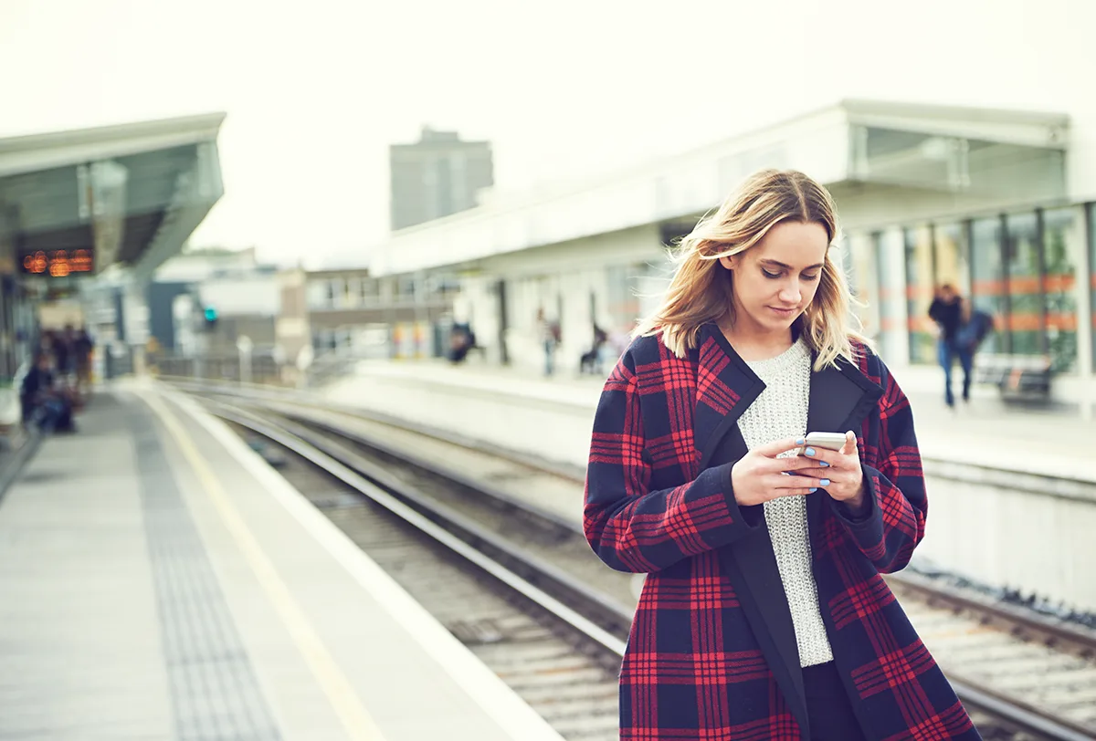 A young white woman with long blonde hair and a red and blue checked coat, standing on a train platform in the daytime, looking at her mobile phone,