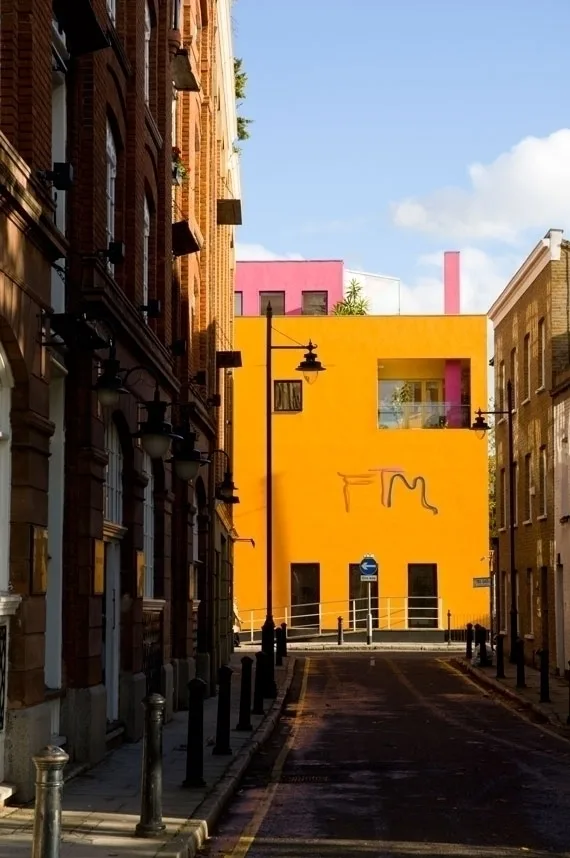 Narrow London street with traditional brick buildings leading to a vibrant yellow and pink modern building under blue sky.