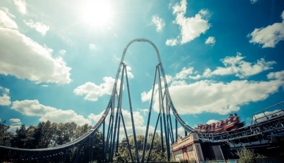 A high, needle-style roller coaster against a blue sky dotted with white clouds. 