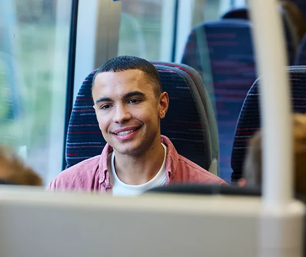 A young man in a pink shirt smiling while seated at a table on a train with other passengers.