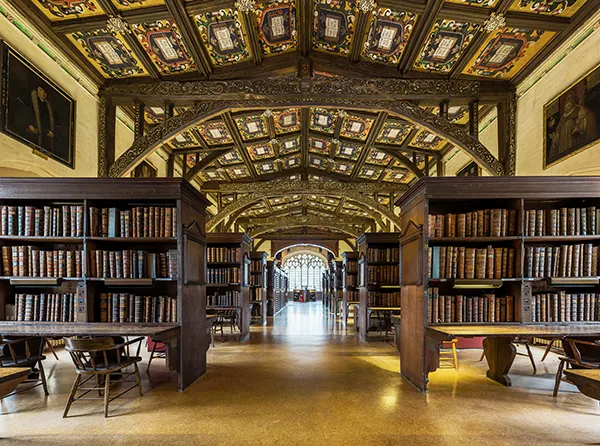 The empty interior of a historic library building with high wooden beams and a decorated ceiling, rows of dark wooden cases hold old books.