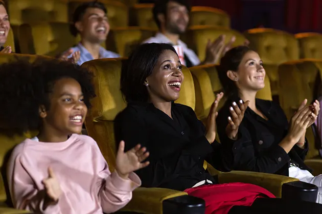 Close up of the faces of audience members in a theatre sitting on yellow chairs and clapping a performance.
