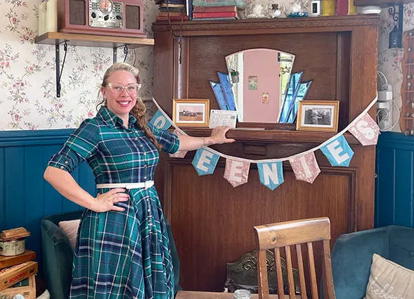 A white woman with blonde hair in a ponytail and cats eye glasses, wearing a blue checked dress. She is standing in a room furnished in vintage 1940s style, in front of pink and blue bunting that spells out QUEENIE'S in white letters. 