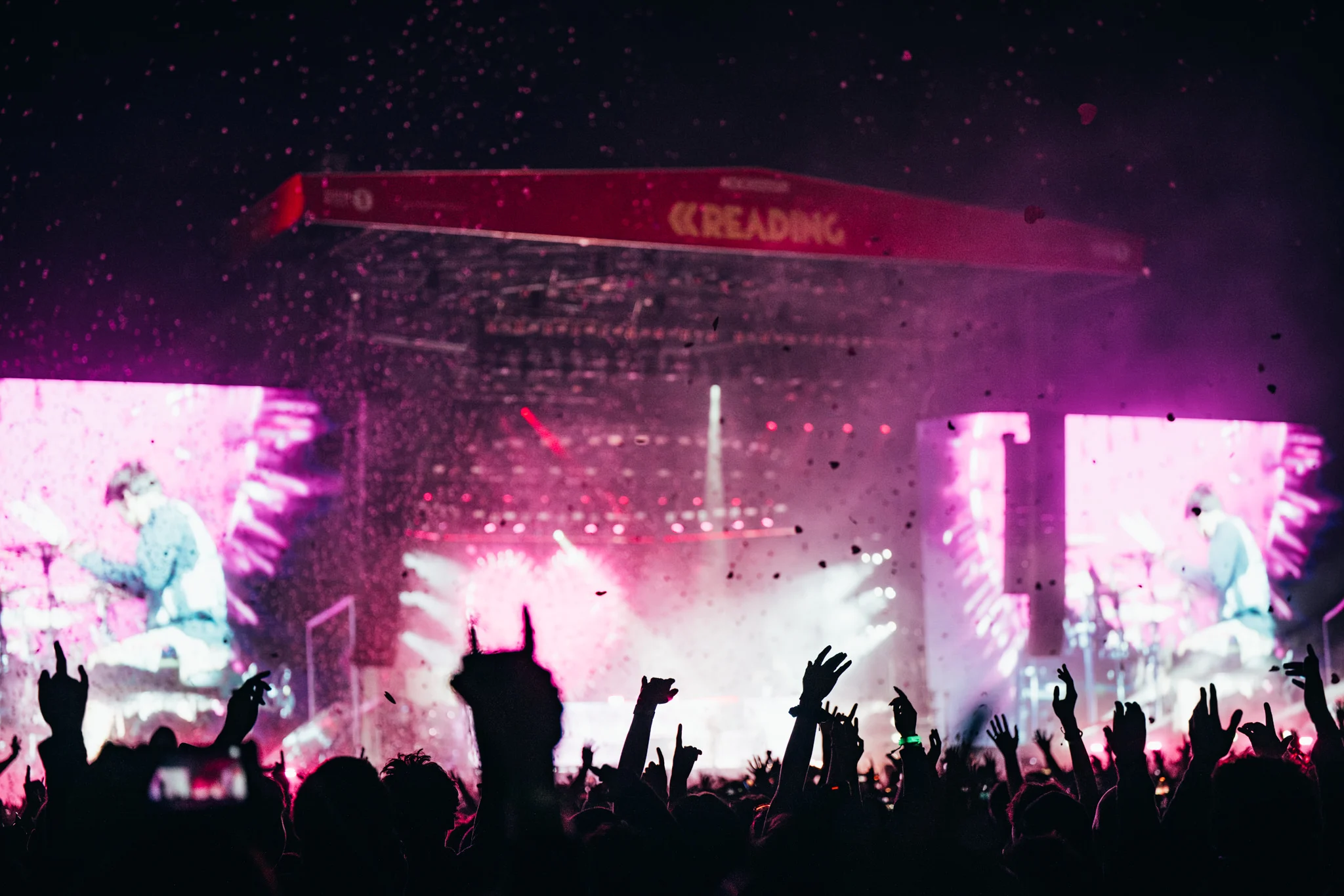 A crowd with raised hands at Reading Festival, with a pink-lit stage, confetti, and large screens displaying a performer.