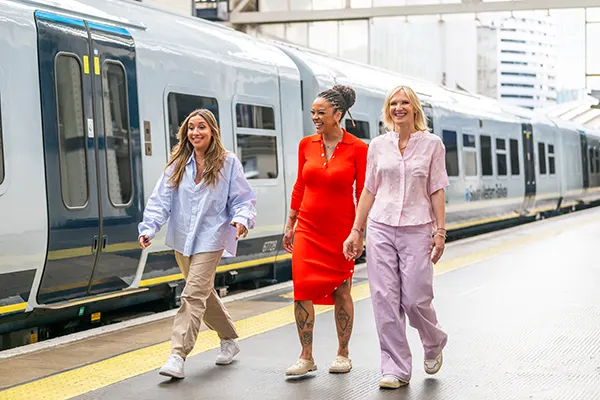 Melanie Blatt, a white woman with long brown hair, Yinka Bokinni, a Black woman with dark braided hair, and Jo Whiley, a white woman with blonde hair, wearing summery clothes and smiling as they walk along a train platform. 