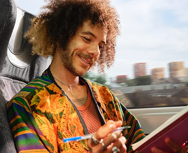A Black man with curly hair and beard, wearing colourful patterned jacket using a tablet while seated on a train with view of a city from the window. 