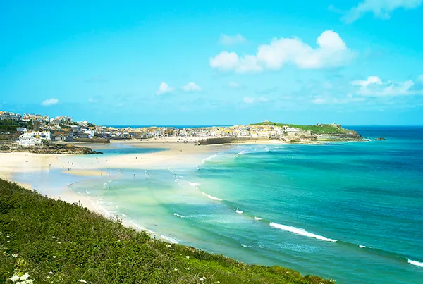 Looking across Carbis Bay in Cornwall on a sunny day, with a sandy beach and bright blue sea and sky, and clifftops in the distance.