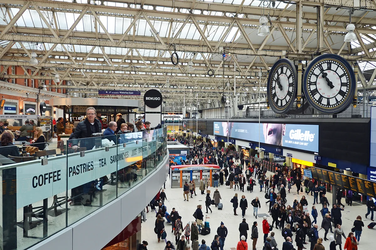 The concourse of London Waterloo station with a large clock hanging from a ceiling comprised of white beans and glass. People are walking about and standing on a balcony overlooking the main area. 