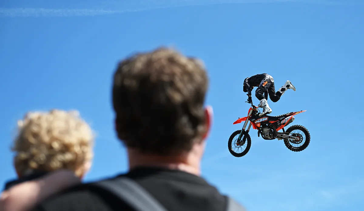 Motocross rider performing aerial stunt on orange dirt bike against blue sky, with spectators watching in foreground.
