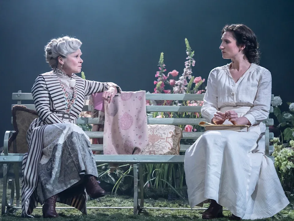 A theatrical production where an older woman with grey hair and a younger woman with dark hair, in Edwardian clothing, are sitting on a bench in a garden