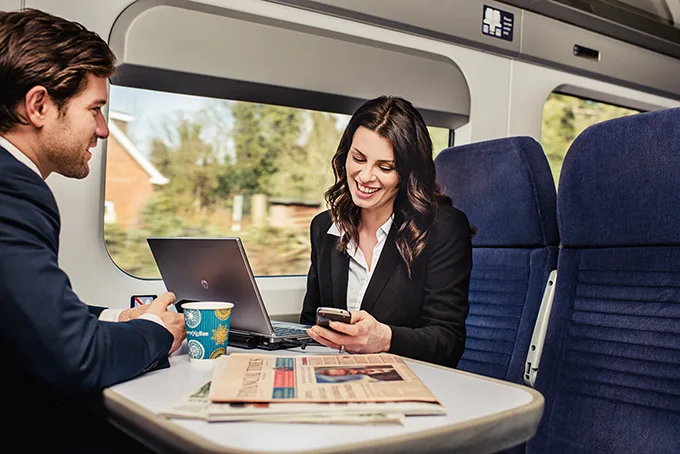 A white man and white woman are working at a table on a train, with newspapers spread out next to a laptop. 