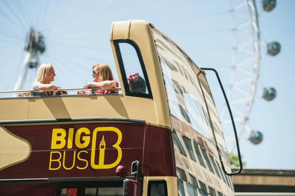 Two women chat to each other on the top deck of a big bus coloured in cream and wine, with the yellow Big Bus logo on the side of the bus. The London Eye sits in the background in front of a blue sky.