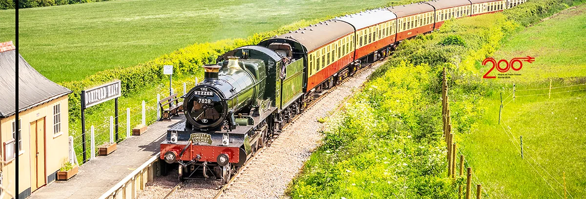 A heritage stream train on a track at a small station with green fields either side. 