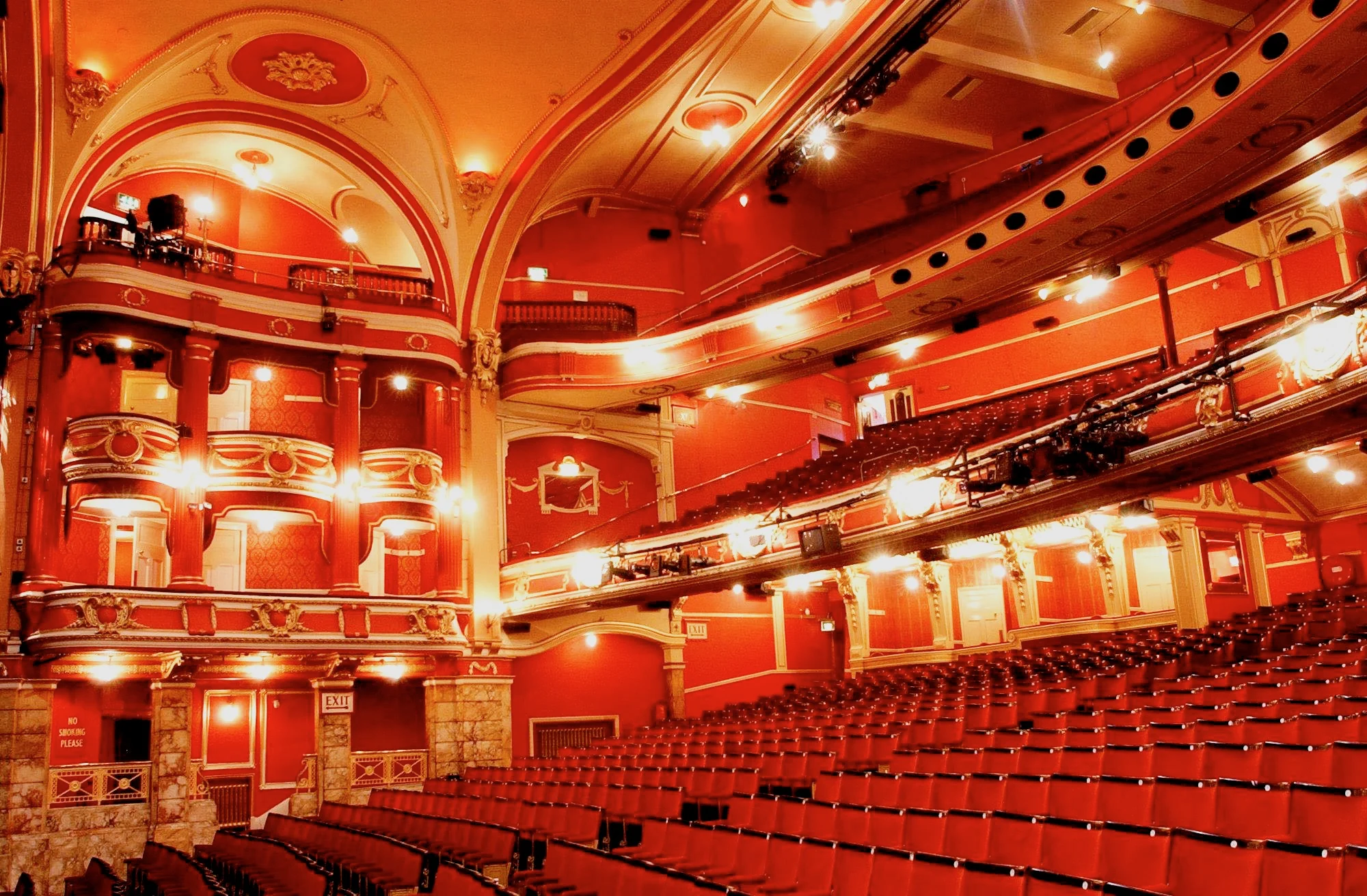 The brightly lit interior of an empty theatre with red seats and decorated boxes and ceiling.