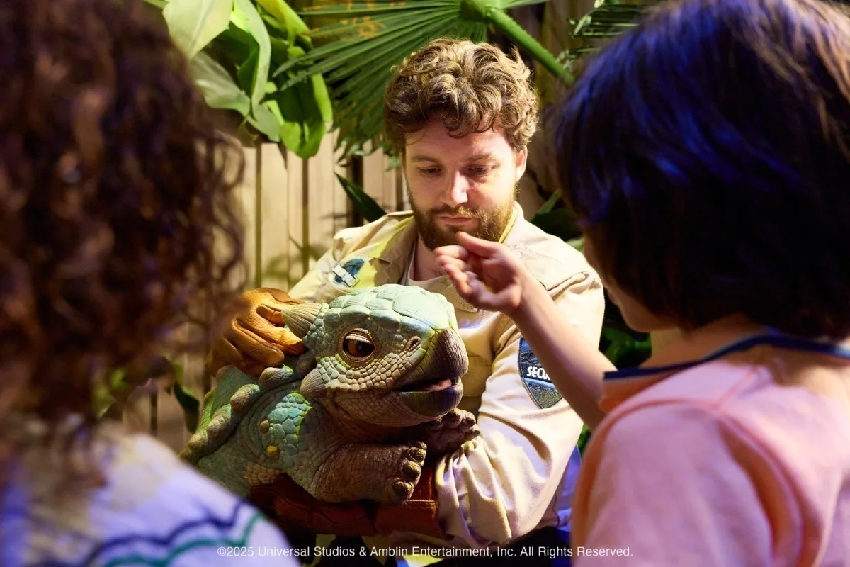 A white male zoo keeper with brown hair and beard, wearing a tan uniform and holding a large green dinosaur puppet while children reach out to touch it in a tropical setting.