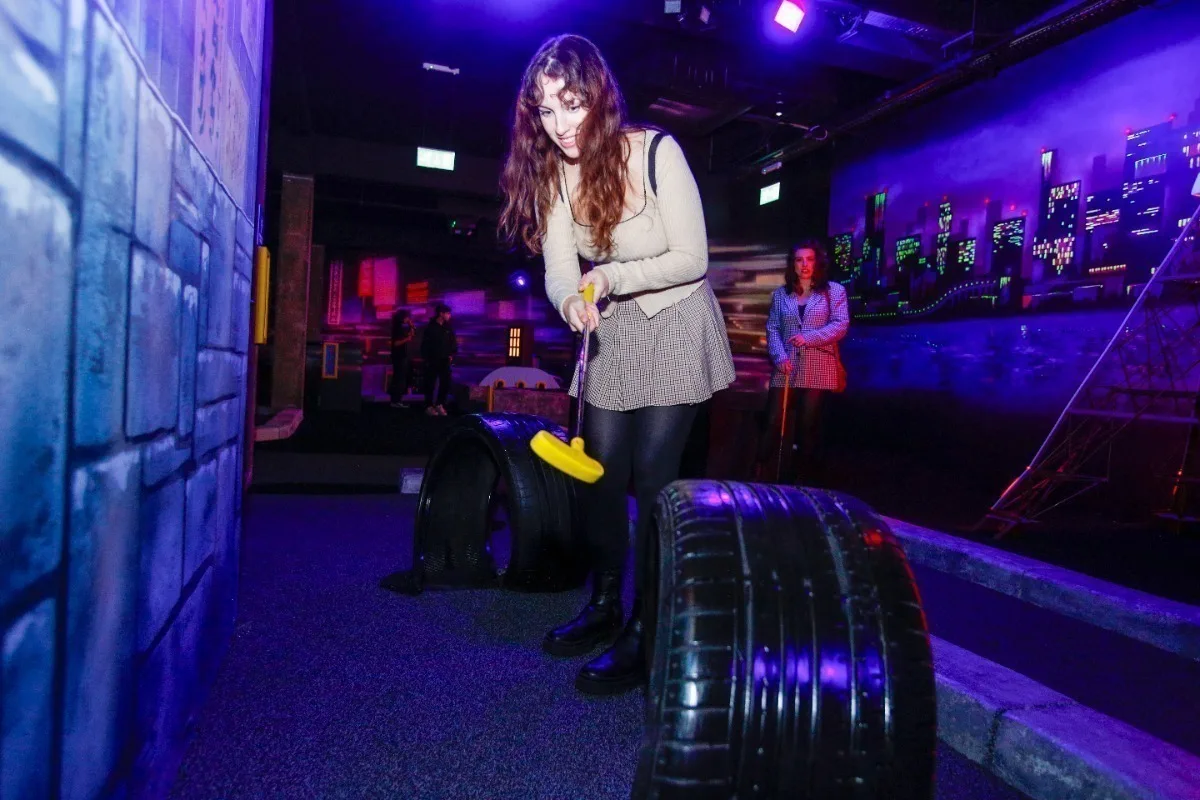 A white woman with long dark hair holding a crazy golf club and standing in a purple-lit room.