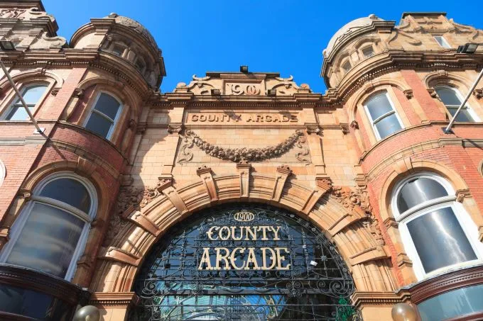 County arcade entrance beneath blue sky. The words county and arcade are are written as a sign in intricate letters on top of glass and dark metal casing. The brick surrounding is orange and cream.