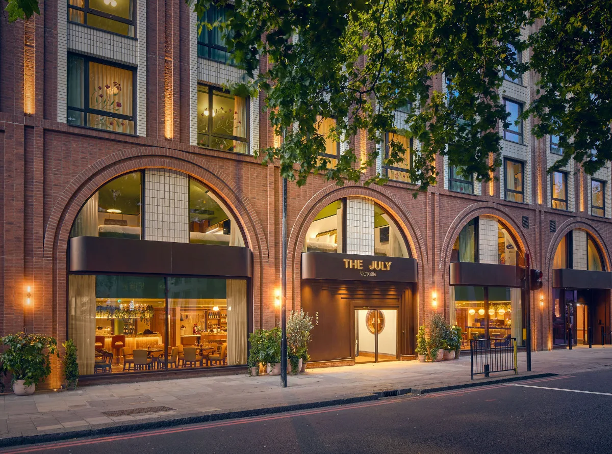 Exterior of The July Victoria hotel at dusk, featuring red brick arches, warm lighting, and a restaurant visible through large windows.