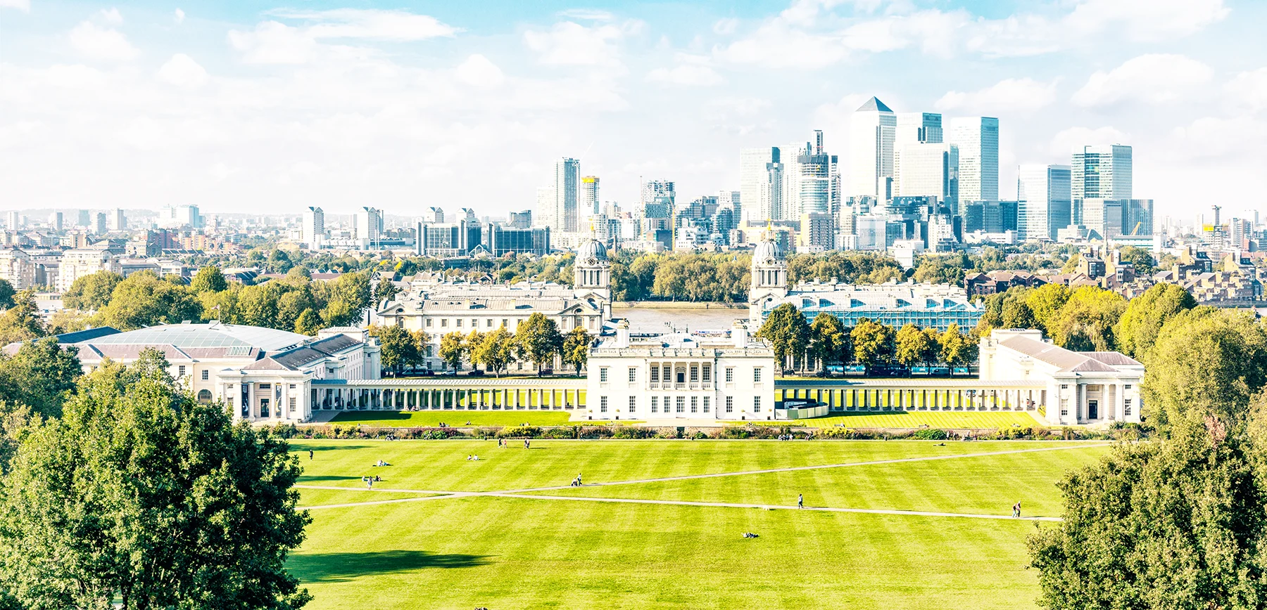 A green park with historic buildings in Greenwich, and the London skyline with several large skyscrapers in Canary Wharf in the background.