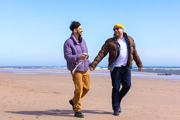Two men walking hand-in-hand on a sandy beach with clear blue sky and sea in the background.